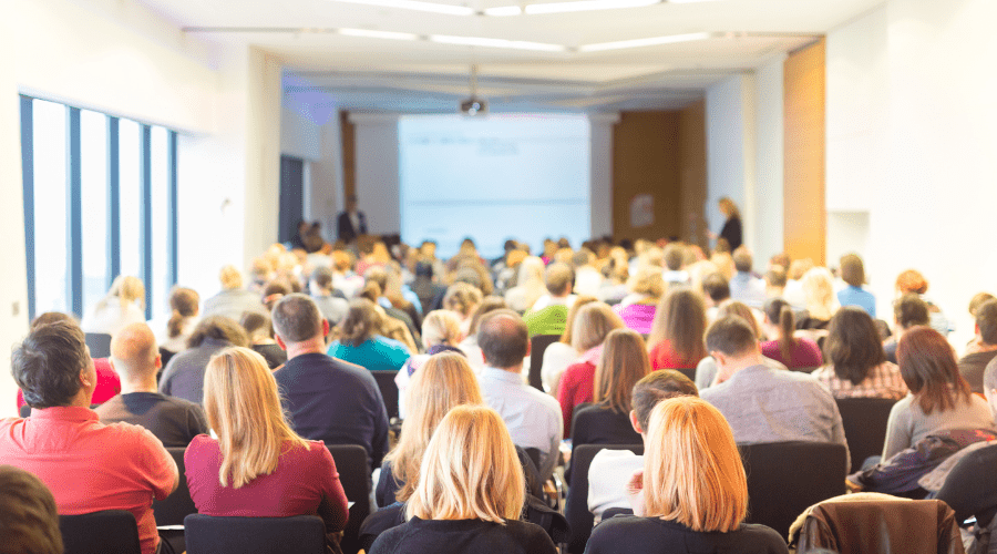 A room full of people listening to a speaker at a coference.